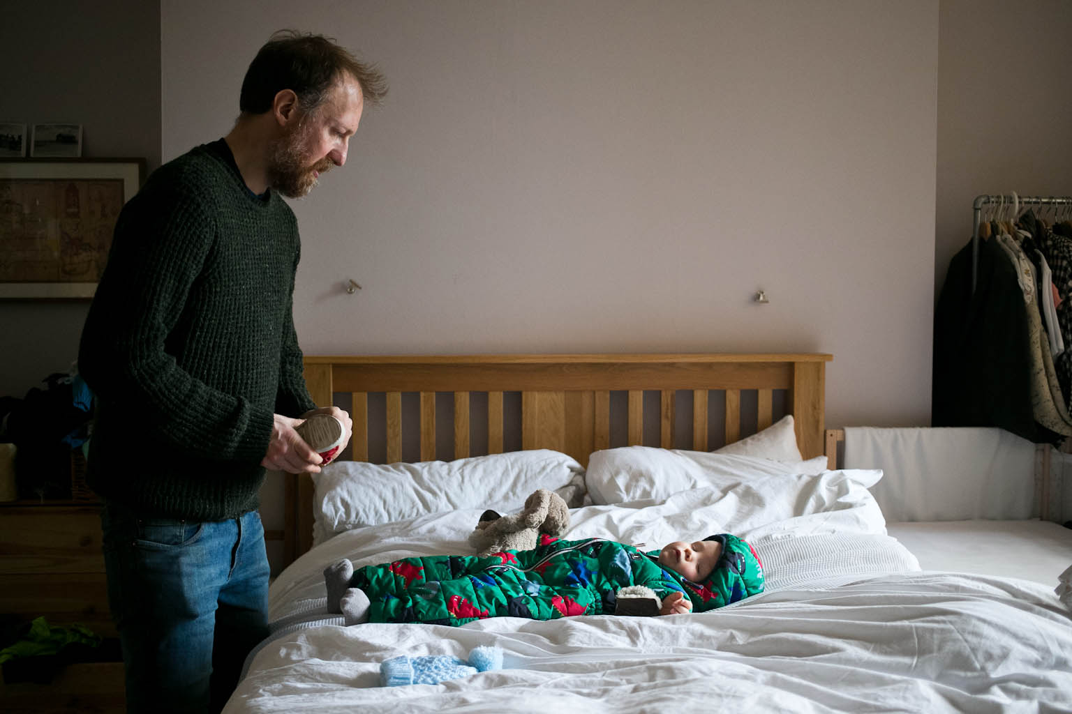 Father holding little shoe looking at sleeping baby boy on bed wearing outdoor suit