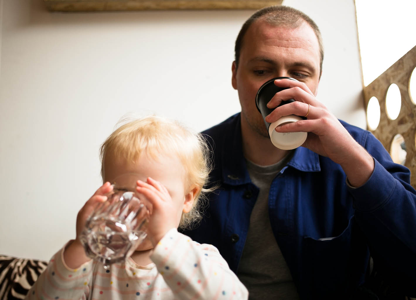 Father drinking takeaway coffee watching young daughter drink a glass of water