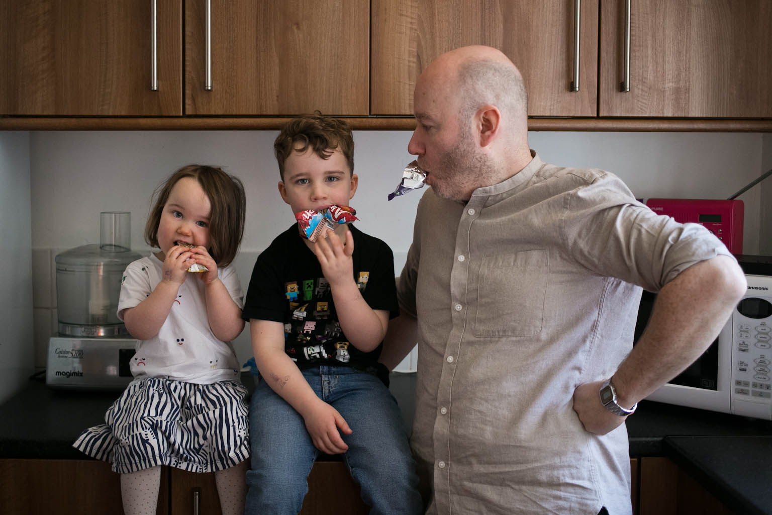 Siblings eating snack bars looking to camera while dad jokes with snack bar in mouth